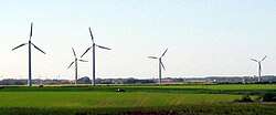 A car drives past 4 wind turbines in a field, with more on the horizon