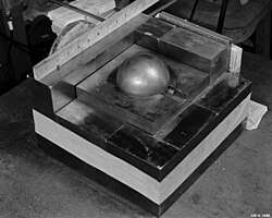 A stack of square metal plates with a side about 10 inches. In the 3-inch hole in the top plate there is a gray metal ball simulating Pu.