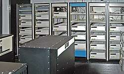 A room with a black box in the foreground and six control cabinets with space for five to six racks each. Most, but not all, of the cabinets are filled with white boxes.